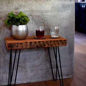 May include: A small, rustic wooden side table with black hairpin legs. The tabletop holds a silver pot with greenery, two glasses, one with red liquid, and one with water. A modern, minimalist design.