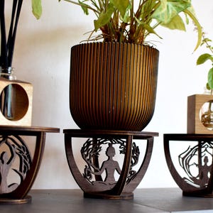 May include: Three wooden plant stands with intricate laser cut designs featuring a stylised human figure. Each stand has a round top and a base with a decorative pattern. The stands are made of dark wood and are displayed on a light wood surface.