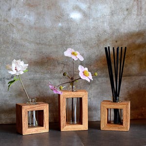 May include: Three wooden frames with glass vials, two containing pink flowers and one with black incense sticks. The frames are on a grey concrete surface.