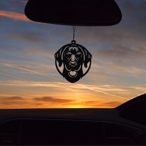 May include: Black metal car charm featuring a dog's head wearing sunglasses. The charm is hanging from a string, silhouetted against a colorful sunset. The dog's face is detailed with cut-out features.