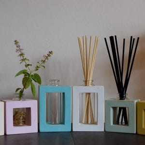 May include: Six colourful, square-shaped essential oil diffusers. The diffusers are in pastel shades of white, pink, blue, and yellow. Some contain reeds, others hold plants or are empty. The diffusers are arranged in a row, creating a decorative display.