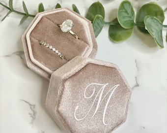 May include: A blush pink velvet ring box with an octagonal shape. The box is open and has a ring inside. The box is monogrammed with the letter "M".