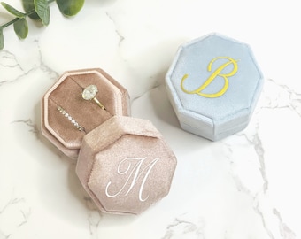 May include: Two velvet ring boxes, one light blue with the letter "B" embroidered in gold, and one blush pink with the letter "M" embroidered in white.