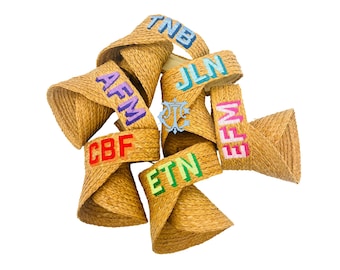 Puede incluir: Una colección de viseras de paja tejida, cada una con iniciales bordadas en varios colores. Las viseras son de color beige natural, con iniciales en azul, morado, rojo, verde y rosa.
