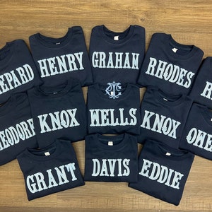 May include: A collection of 12 navy blue t-shirts with white embroidered names, including Shepard, Henry, Graham, Rhodes, Knox, Wells, Owen, Theodore, Grant, Davis, Eddie, and Riv.