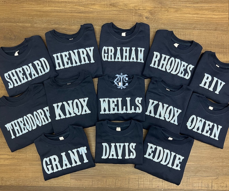May include: A collection of 12 navy blue t-shirts with white embroidered names, including Shepard, Henry, Graham, Rhodes, Knox, Wells, Owen, Theodore, Grant, Davis, Eddie, and Riv.
