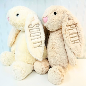 May include: Two plush bunny toys, one cream colored and one light brown, are sitting side by side. The cream colored bunny has the name "Scotty" embroidered on its ear, and the light brown bunny has the name "Piper" embroidered on its ear.