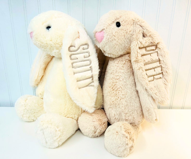 May include: Two plush bunny toys, one cream colored and one light brown, are sitting side by side. The cream colored bunny has the name "Scotty" embroidered on its ear, and the light brown bunny has the name "Piper" embroidered on its ear.
