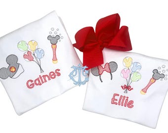 Puede incluir: Dos camisetas blancas con bordados de orejas de Mickey y Minnie Mouse, globos de colores y varitas de burbujas. Una camiseta muestra el nombre Gaines en rojo, mientras que la otra muestra el nombre Ellie en rojo. Se incluye un lazo rojo grande.