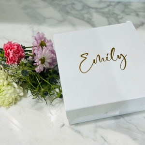 May include: A white gift box with a gold inscription that reads "Emily". The box is sitting on a white marble surface with a bouquet of pink, white, and purple flowers.
