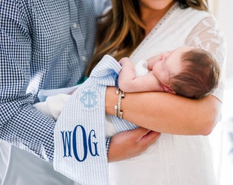Puede incluir: Una manta de bebé a rayas azules y blancas con el texto "The Classy Llama" y "WOG" bordado. Un bebé está siendo sostenido por un padre.