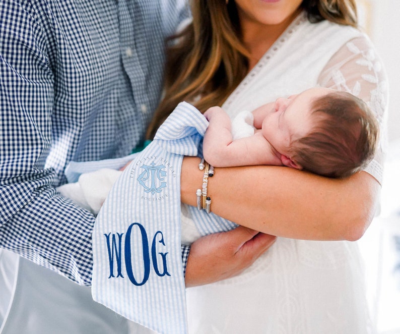 May include: A blue and white striped baby blanket with the text "The Classy Llama" and "WOG" embroidered on it. A baby is being held by a parent.
