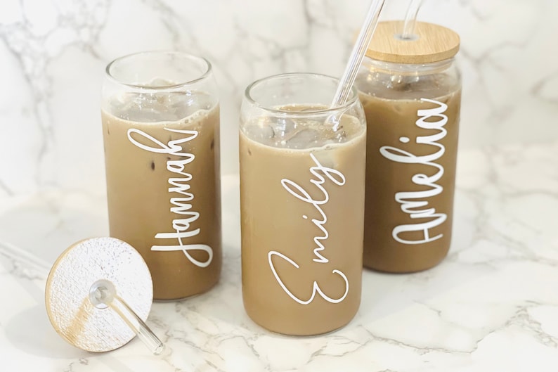 May include: Three glass can-shaped cups with lids and straws filled with iced coffee. The cups are personalized with the names "Hannah", "Emily", and "Amelia" in white lettering.