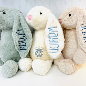 May include: Three plush bunny toys with embroidered names on their ears. The bunnies are in shades of green, cream, and beige. The cream bunny has a logo on its chest that reads "The Classy Thread Boutique".
