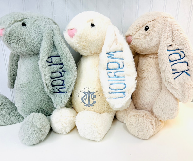 May include: Three plush bunny toys with embroidered names on their ears. The bunnies are in shades of green, cream, and beige. The cream bunny has a logo on its chest that reads "The Classy Thread Boutique".