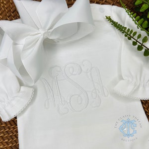 May include: White baby onesie with a white embroidered monogram "MSI" on the front. The onesie has a ruffled trim around the sleeves and neckline. The onesie is laying on a woven basket with a white bow on top. The bottom right corner of the image has a logo that says "The Classy Thread Boutique".