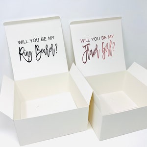 May include: Two white gift boxes with lids open. The box on the left has the text "Will you be my Ring Bearer?" printed on the lid. The box on the right has the text "Will you be my Flower Girl?" printed on the lid.