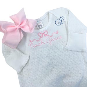 May include: White baby sleeper with a pink bow and the embroidered name "Noah Grace". The sleeper has long sleeves with folded cuffs and a textured pattern. A large pink bow is attached to the shoulder.