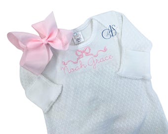 May include: White baby sleepsuit with a pink bow and the embroidered name "Noah Grace". The sleepsuit has long sleeves with folded cuffs and a textured pattern. A large pink bow is attached to the shoulder.
