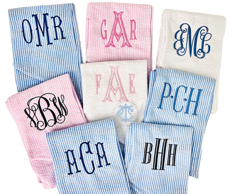 May include: A collection of eight personalized towels with different embroidered monograms. The towels are in various colors, including pink, white, and blue. The monograms are in different styles, including script and block letters.