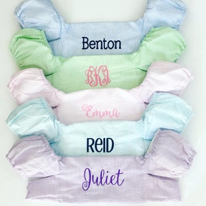 May include: Five different colored, striped, fabric baby bibs with embroidered names. The names are Benton, BKA, Emma, Reid, and Juliet.