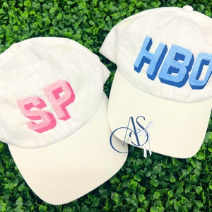 May include: Two white baseball caps with embroidered letters. The cap on the left has the letters "SP" in pink. The cap on the right has the letters "HBO" in blue.