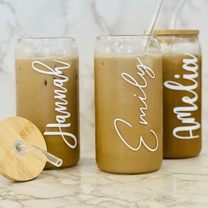May include: Three clear glass can-shaped cups with lids and straws. The cups are filled with a brown liquid and have white lettering on the front. The lettering reads "Hannah", "Emily", and "Amelia".