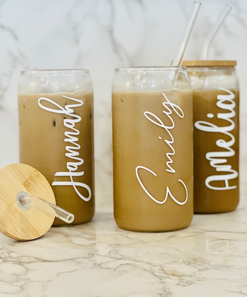 May include: Three clear glass can-shaped cups with lids and straws. The cups are filled with a brown liquid and have white lettering on the front. The lettering reads "Hannah", "Emily", and "Amelia".