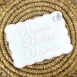 Puede incluir: Un parche de tela blanco con un borde bordado rosa y el texto "Alexa Elliette Alanis" en letra cursiva rosa.