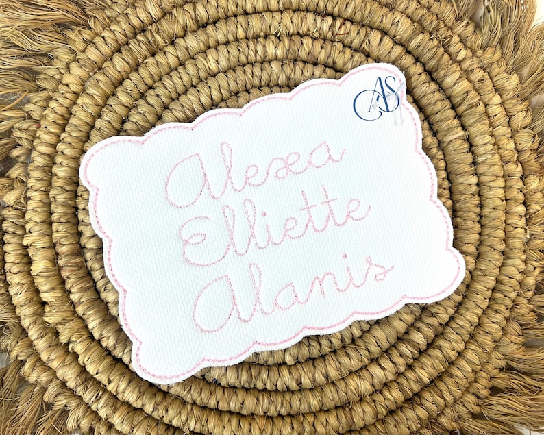 Puede incluir: Un parche de tela blanco con un borde bordado rosa y el texto "Alexa Elliette Alanis" en letra cursiva rosa.