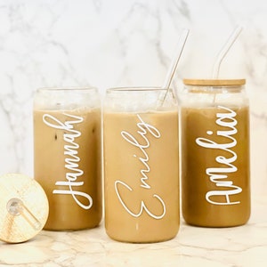May include: Three glass beverage containers with lids and straws. The containers are filled with a brown liquid and have personalized names on them: "Hannah", "Emily", and "Amelia".