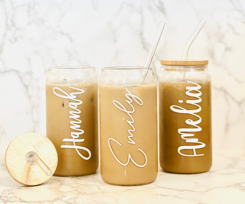 May include: Three glass beverage containers with lids and straws. The containers are filled with a brown liquid and have personalized names on them: "Hannah", "Emily", and "Amelia".