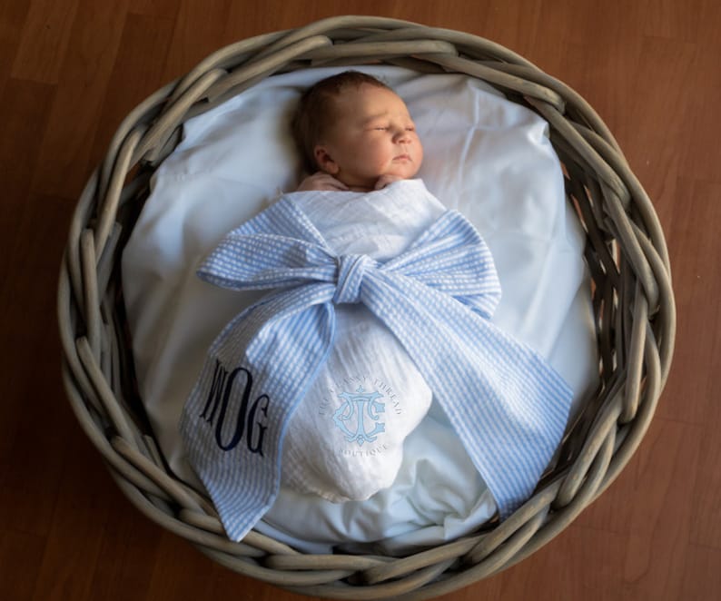 May include: A newborn baby is sleeping in a wicker basket, wrapped in a blue and white striped blanket with a bow. The blanket is monogrammed with the letter "J" and the word "DOG".