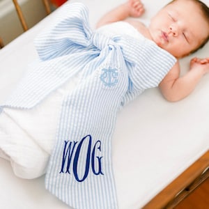 May include: A baby swaddled in a blue and white striped swaddle with a monogrammed bow. The swaddle is tied in a bow at the top and has the text "The Classy  Thread" and an anchor symbol on the front. The monogram on the bow is "WOG".