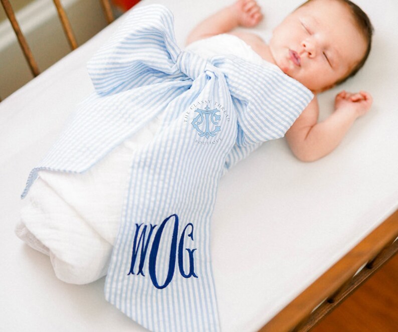 May include: A baby swaddled in a blue and white striped swaddle with a monogrammed bow. The swaddle is tied in a bow at the top and has the text "The Classy  Thread" and an anchor symbol on the front. The monogram on the bow is "WOG".
