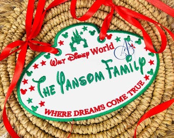 Puede incluir: Una etiqueta de nombre ovalada blanca con texto bordado en verde que dice "Walt Disney World The Pansom Family Where Dreams Come True". La etiqueta está decorada con estrellas rojas y cabezas de Mickey Mouse. La etiqueta está unida a una cinta roja.