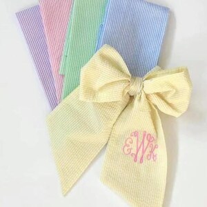 May include: A yellow gingham bow with a pink monogram "EWK" on the center. The bow is made from a fabric with a subtle striped pattern. The bow is surrounded by four pieces of fabric in different colors: light blue, green, pink, and purple. Each fabric has a striped pattern.