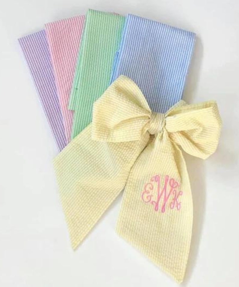 May include: A yellow gingham bow with a pink monogram "EWK" on the center. The bow is made from a fabric with a subtle striped pattern. The bow is surrounded by four pieces of fabric in different colors: light blue, green, pink, and purple. Each fabric has a striped pattern.