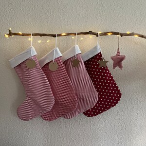 Könnte beinhalten: Vier rot-weiß gestreifte Weihnachtsstrümpfe mit weißen Bündchen und braunen Etiketten hängen an einem Ast mit weißen Lichterketten. Ein rot-weiß gestreifter Sternenschmuck hängt am Ast.