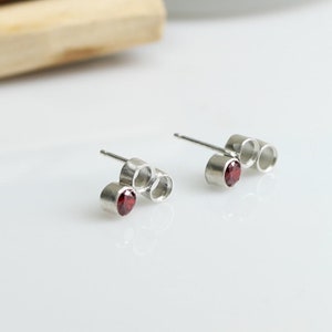 May include: Silver stud earrings, each with a small, round, red gemstone. The earrings have a minimalist design with a post and three connected circular elements. A bundle of wood sticks tied with a maroon ribbon is in the background.