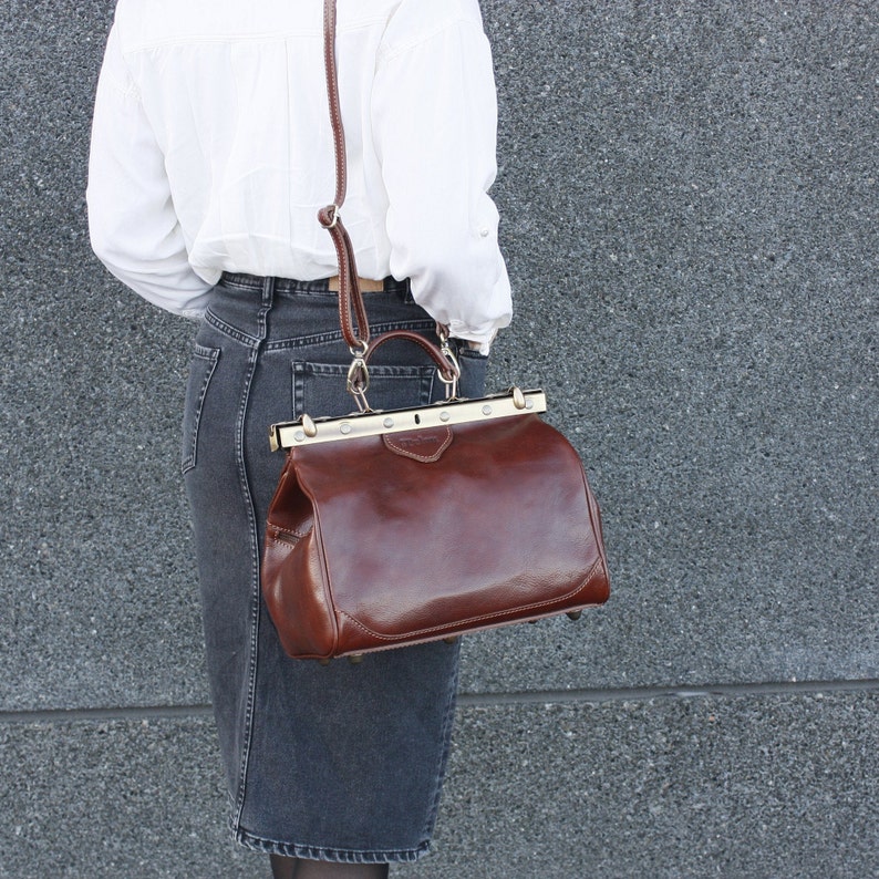 May include: A brown leather satchel bag with a top handle and a shoulder strap. The bag has a rectangular shape with a metal clasp closure and a structured design. The bag is worn over the shoulder.