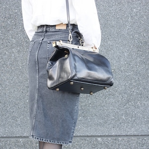 May include: Black leather handbag with a structured design, featuring a top handle and a shoulder strap. The bag has a metal frame and small metal feet on the bottom. The bag is held by a person wearing a white blouse and a denim skirt.