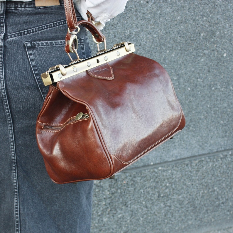 May include: A brown leather satchel with a structured design, featuring a top handle and a metal clasp closure. The bag has a zipper pocket on the front and a shoulder strap. The bag is approximately 12 inches wide.