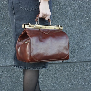 May include: A brown leather handbag with a brass clasp and handle is held by a person. The bag has a structured shape, a zipper pocket, and a glossy finish. The bag is a classic style, suitable for everyday use.