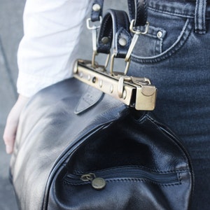 May include: Black leather duffel bag with a brass-colored clasp and zipper. The bag has a rounded shape and a top handle with black leather straps. The zipper pull and clasp are a matching brass color. The bag is attached to a black leather strap.