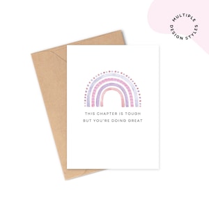 Peut inclure: Une carte de vœux blanche avec un arc-en-ciel aquarelle dans les tons violets et roses. Elle est accompagnée d'une enveloppe marron. Le texte sur la carte se lit : "THIS CHAPTER IS TOUGH BUT YOU'RE DOING GREAT."