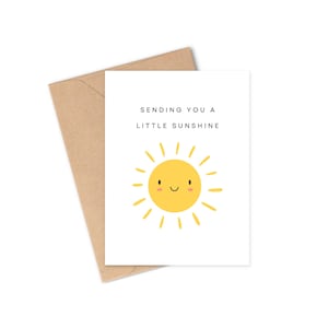 Op de afbeelding: Een witte wenskaart met de tekst "Sending You A Little Sunshine" en een gele, lachende zon-illustratie. De kaart wordt vergezeld door een bruine envelop.