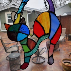 May include: A colorful stained glass cat with a curved back and tail, suspended by a chain. The cat features a patchwork design of various colors, including blue, red, yellow, green, and purple. The artwork is displayed in front of a window.