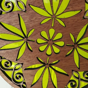 Puede incluir: Un adorno redondo de madera con un diseño verde de hojas de cannabis y una flor estilizada rellenas de resina. El adorno tiene un fondo de madera marrón.