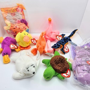 May include: A collection of Ty Beanie Baby plush toys including a purple platypus, yellow duck, orange fish, pink flamingo, blue and black lizard, white seal, and green turtle, with two items still sealed in clear plastic packaging with printed text.